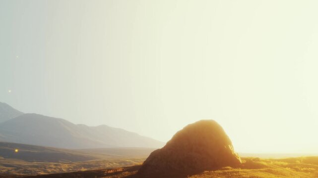 The sun sets behind mountains, casting light on a large rock formation. The sky displays warm tones as the landscape becomes shadowed.
