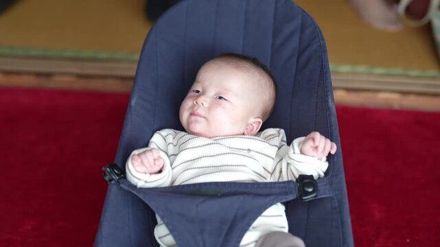 A top-down view captures an alert infant stretching and moving both arms in a supportive bouncer.