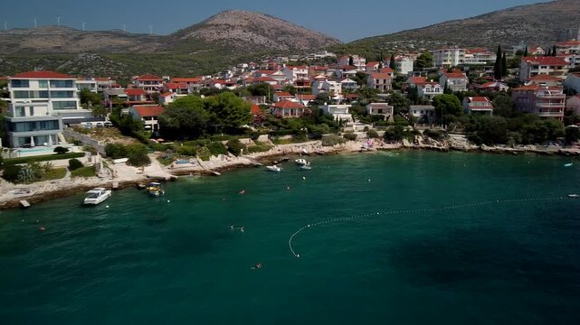 Seget Vranjica, Trogir, Croatia homes line turquoise Adriatic shoreline below hills, aerial dynamic pan right across coastal buildings
