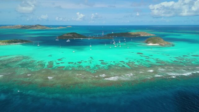 Aerial approach descends from high altitude toward Tobago Cays, revealing Petit Rameau, Baradal, Jamesby and Petit Tabac islands scattered with sailboats across turquoise atoll sea
