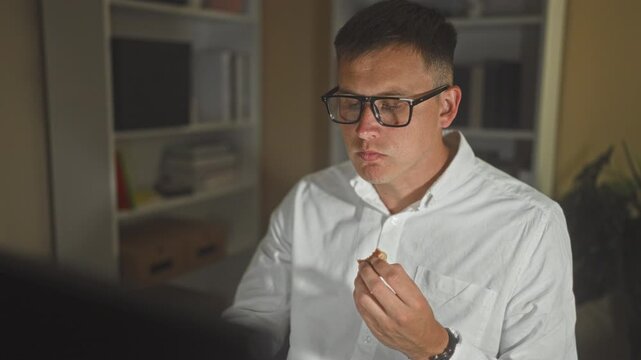 Man wearing glasses holds a cookie and chews while looking at a computer monitor at a desk in a building; late night focus.