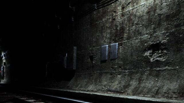 A dark subway tunnel is shown with railway tracks running along the ground. The walls are rough and worn, with visible signs of age and use. There are no people in sight.