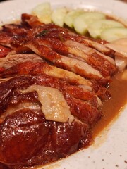 Obraz premium A close-up shot of succulent sliced roasted duck topped with savory gravy sauce, accompanied by sliced fresh cucumbers and pickled ginger on a white plate.