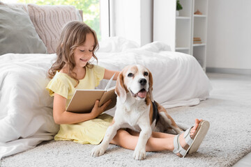 Fototapeta premium Cute little girl with Beagle dog reading book on floor in bedroom