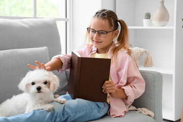 Fototapeta premium Cute little happy girl with Maltese dog reading book on sofa at home