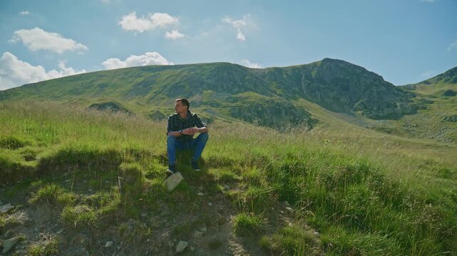 Man sits on grassy hill while looking at mountain landscape under bright sky with clouds