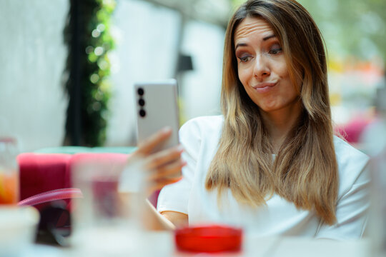 Skeptical Young Woman Reading Message On Phone or Making Selfies. Doubtful female customer holding mobile smartphone inside cafe looking worried about digital text