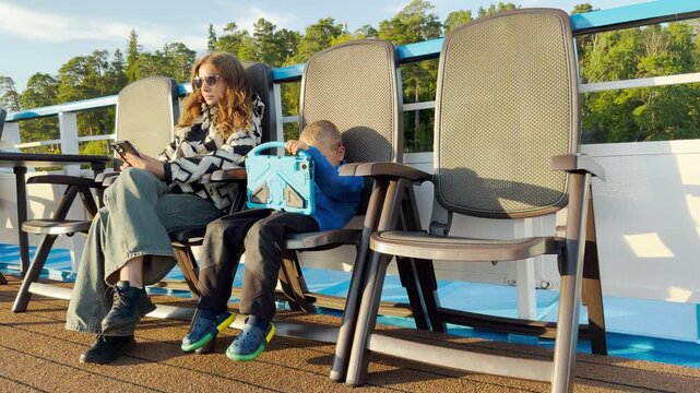 A mother and her young son relax on deck chairs while looking at their devices aboard a cruise ship. They feel calm and content on a sunny trip across Lake Ladoga.