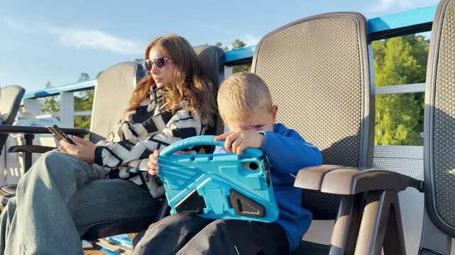 A mother and her young boy sit on deck chairs while using their screen devices on a cruise ship. They feel calm and focused while cruising along scenic Lake Ladoga.