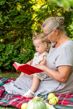 Grandmother Reading Red Book With Granddaughter During Garden Picnic