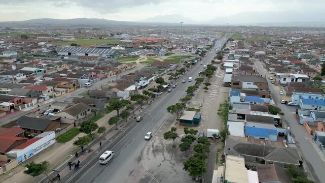 Vehicles move along a central avenue lined with low-rise homes and small shops in Langa Township under cloudy skies.