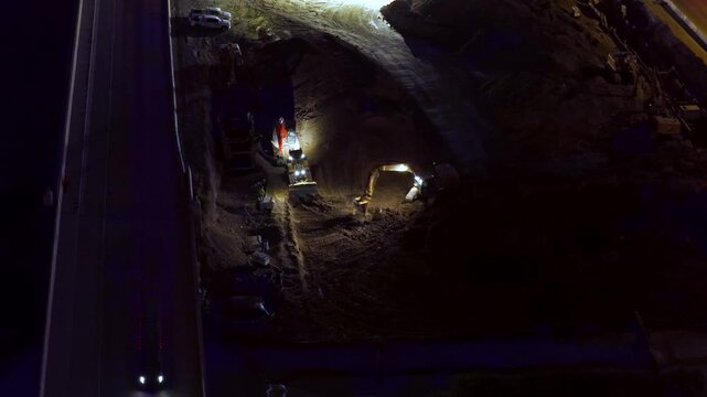 Static drone aerial shows illuminated heavy machinery building a freeway along a frontage road at night, with glowing headlights, dark surroundings, strong contrasts, and visible worksite activity.