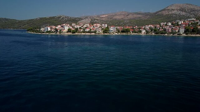Seget Vranjica, Trogir, Croatia coastal town stretches beside blue Adriatic Sea, aerial establishing dolly with wind turbines on ridge above deep blue water