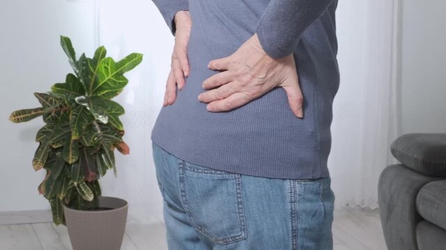 Back pain. Middle aged woman holding her lower back and massaging it with her hands at home. Close-up, rear view, side view. Lumbago, sciatica, lower back pain, lumbago in the back, hernia, protrusion