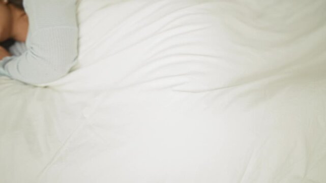 Woman sleeping on bed with hands near face, covered by white duvet, pearl earring visible in a studio setting; serenity.
