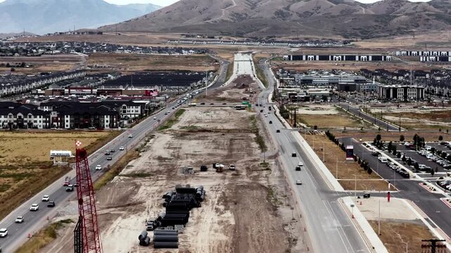 Large-scale freeway construction between active frontage roads, featuring a crane drilling rig, stacks of wastewater pipes, and a bustling urban suburb - pullback aerial reveal