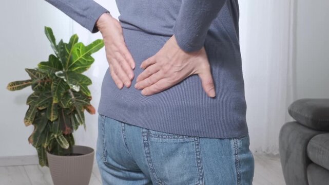 Back pain. Middle aged woman holding her lower back and massaging it with her hands at home. Close-up, rear view, side view. Lumbago, sciatica, lower back pain, lumbago in the back, hernia, protrusion