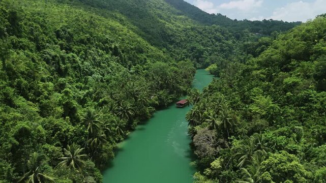 Drone pulls back while tracking a Loboc River cruise boat traveling downstream through green water and forested hills in Bohol