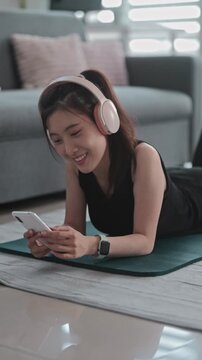 Asian woman in sports clothing using smartphone with headphones at home