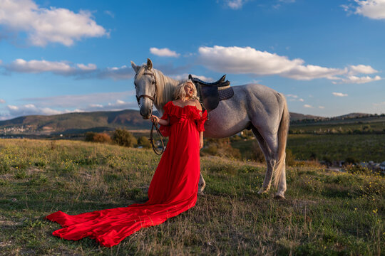 Woman horse red dress majestic blonde model standing in grassy field at sunset