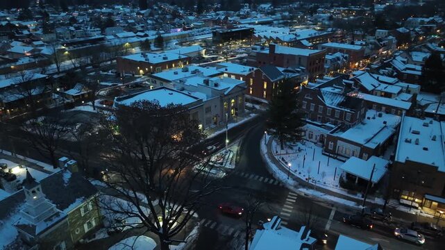Aerial view of a charming small town blanketed in snow, illuminated by streetlights and car headlights during the early evening hours.