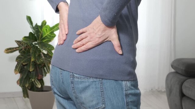 Back pain. Middle aged woman holding her lower back and massaging it with her hands at home. Close-up, rear view, side view. Lumbago, sciatica, lower back pain, lumbago in the back, hernia, protrusion