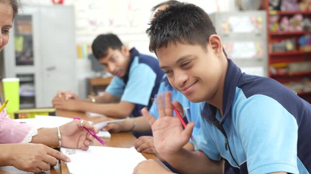 Indian disabled student with down syndrome smiling and waving at the camera while sitting with a teacher in a special needs classroom, representing inclusive education and learning