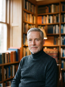 Older man in late sixties with cropped grey hair in fine dark merino roll neck in home library with blurred bookshelves in warm light generative AI