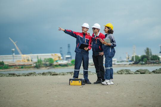 Three marine engineers pointing toward industrial port while discussing vessel inspection, maintenance planning and shipyard operations near shoreline.
