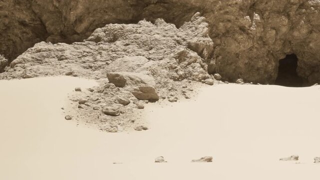 A large rock formation sits in a sandy area near a cave entrance in a desert. The sun shines down on the scene, highlighting the textures of the sand and rocks.