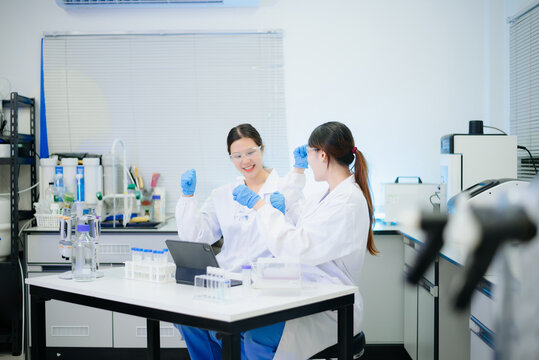 Two Asian female scientists working with modern equipment and a digital tablet in a clean, professional biotechnology laboratory.