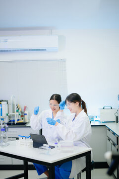 Two Asian female scientists working with modern equipment and a digital tablet in a clean, professional biotechnology laboratory.