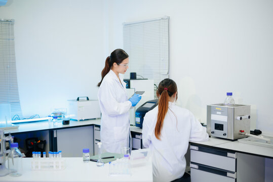 Two Asian female scientists working with modern equipment and a digital tablet in a clean, professional biotechnology laboratory.