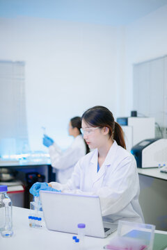 Female scientist analyzing data on laptop with chemical test tube samples in a modern biotechnology laboratory.