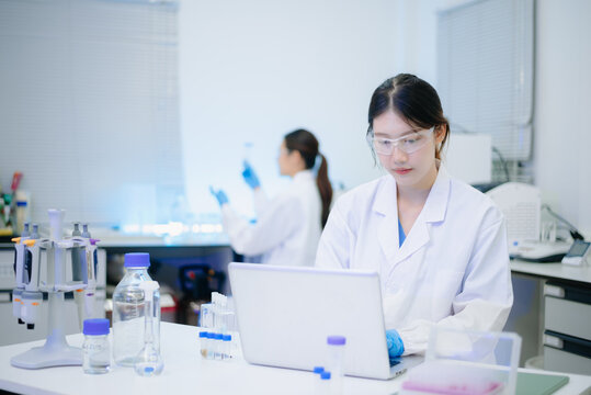 Female scientist analyzing data on laptop with chemical test tube samples in a modern biotechnology laboratory.