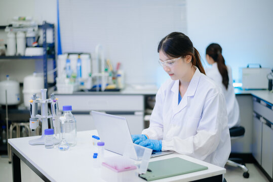 Female scientist analyzing data on laptop with chemical test tube samples in a modern biotechnology laboratory.