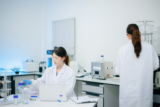 Two Asian female scientists collaborating with a micropipette and laptop computer during research in biotechnology laboratory.