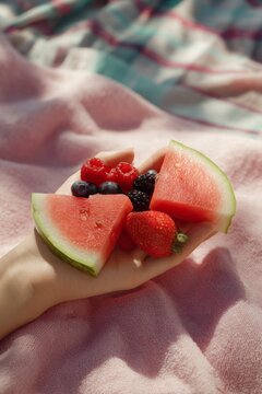 concept of summer relaxation. healthy Gen Z lifestyle. Hand holding assorted fresh fruits including watermelon, strawberries, blueberries, and raspberries on a pink blanket outdoors