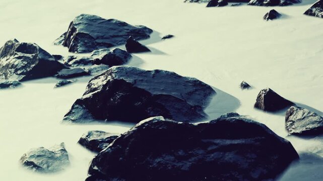 Large rocks are scattered on a beach during low tide. Water covers parts of the sand, reflecting sunlight. Waves gently lap around the rocks while the sky is clear.