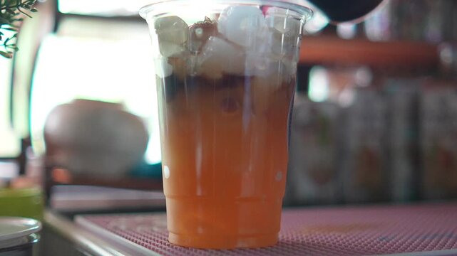 4K Iced coffee layered with orange juice in clear plastic cup filled with ice cubes on cafe counter table for refreshing summer drink concept menu.