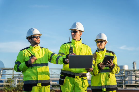 Oil refinery engineers team with laptop and tablet working outdoors at industrial plant, petroleum plant workers in safety gear discussing project with computer and digital devices
