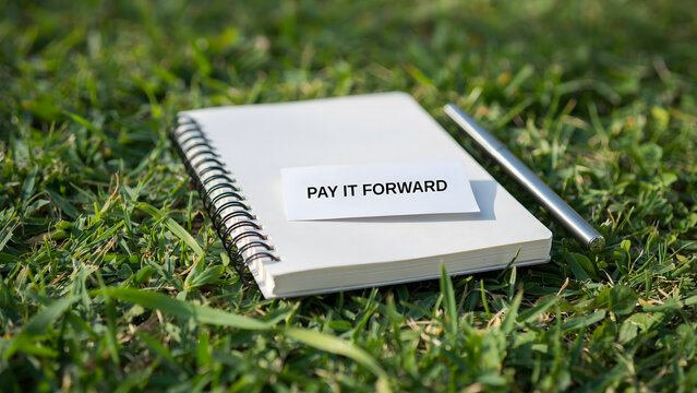 A spiral notebook with a pay it forward sign resting on green grass outdoors