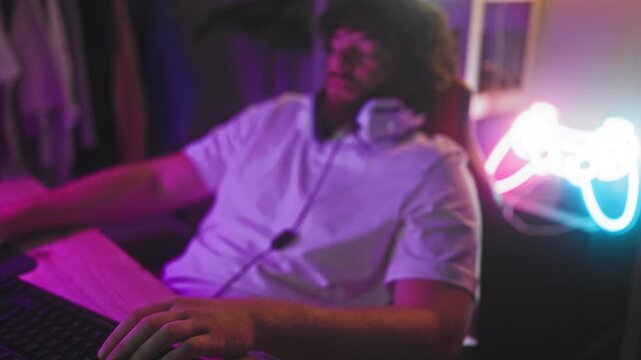 Man with headset leaning back with eyes closed, one hand on keyboard beside neon controller at studio; fatigue.