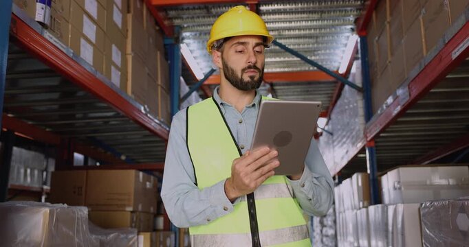 Serious Latin logistic professional man in protective hardhat inspecting storage in hardhat, using shipping app on tablet pc, looking at stacked boxes, packages on metal shelves in warehouse
