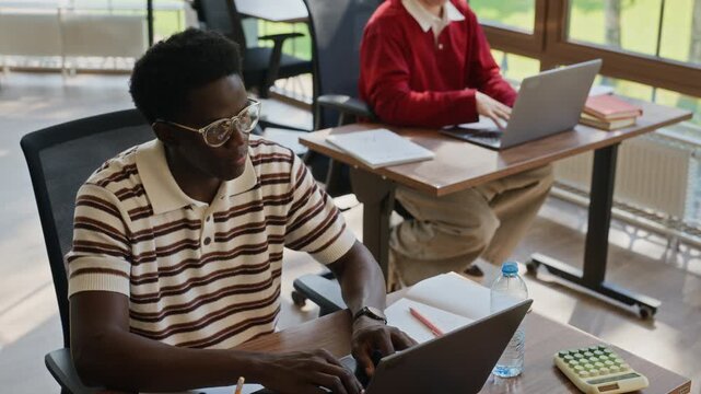 Young adult African American man with glasses typing on laptop while Caucasian colleague works in background. Clip is perfect for corporate training, higher education, or co-working space promos