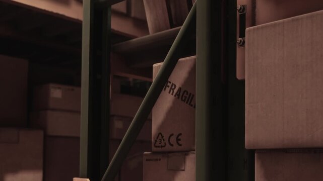 Workers move around boxes in a warehouse. The space is filled with stacked boxes marked as fragile. Safety signs are placed near the aisle. Light comes from the ceiling.