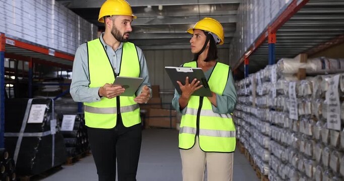 Serious male Latin warehouse worker showing factory space with shelves and boxes to female inspector. of logistic coworkers in hard hats walking in storage, talking, discussing inventory