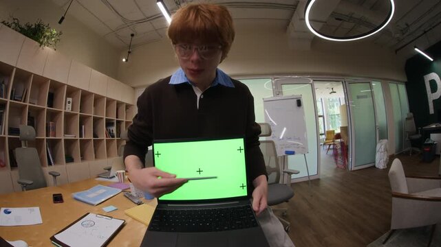 Portrait of young adult Caucasian man presenting laptop with green screen to multiethnic coworkers in office, fisheye view. Clip is perfect for tech startup ads and software promos