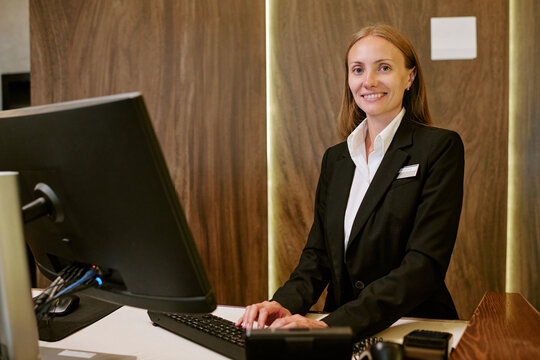 Portrait of smiling Caucasian young adult woman working as hotel receptionist at front desk using computer. Image represents hospitality service and is ideal for tourism marketing