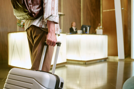 Cropped shot of young adult man holding suitcase handle while walking toward hotel reception desk, representing business travel and hospitality services, low angle view